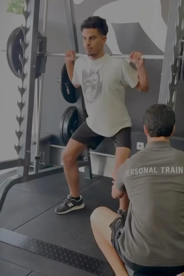 Cliente a fazer agachamento na máquina Smith acompanhado pelo personal trainer