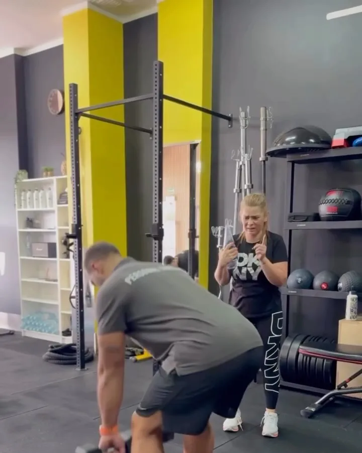 Interior do estúdio com colunas amarelas, onde um personal trainer demonstra um exercício com halter